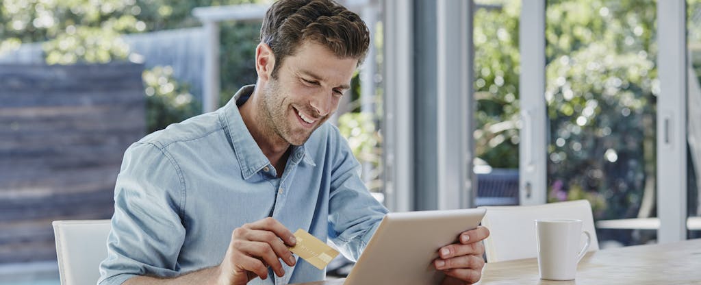 Smiling male in a sunny room, looking at his tablet with a credit card in hand