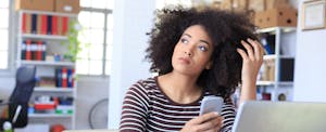 Pensive young woman holding smart phone at workplace. Looking away. As background shelves with boxes and folders, and tall windows with plant, bike and seat. On desk note book, tools, laptop, eyeglasses.