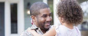 Dad holds his daughter, smiling, upon his return home from military assignment