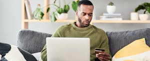 Young man sitting on sofa at home with laptop and holding a credit card