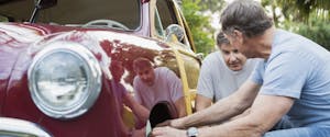Two older men kneeling in front of the wheel of a red vintage car