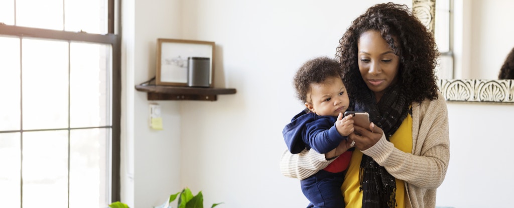 Woman standing in her living room, holding her baby and reading on her phone