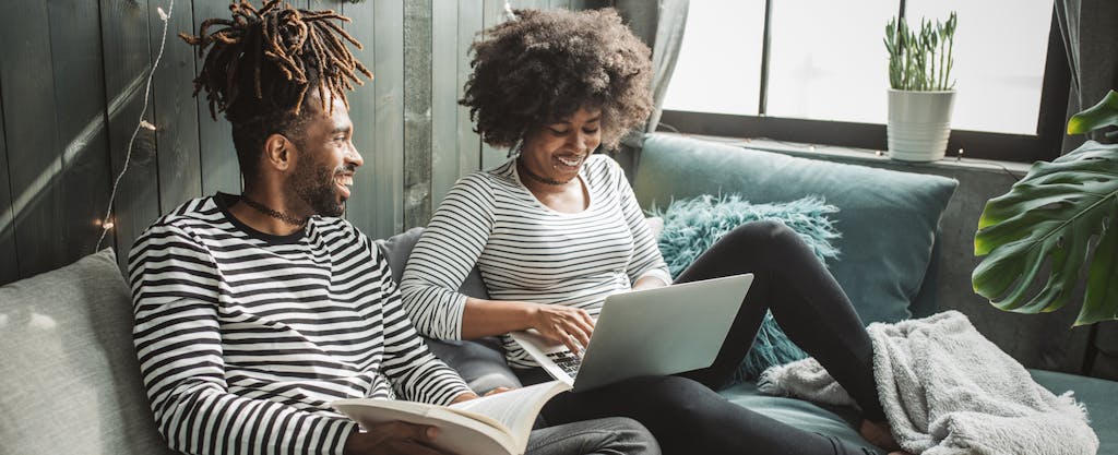 Young couple sitting on couch, reading book and using laptop