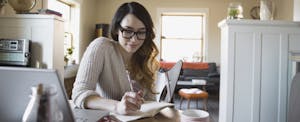 Woman sitting at her kitchen table with her laptop open, writing in a journal