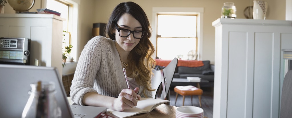 Woman sitting at her kitchen table with her laptop open, writing in a journal