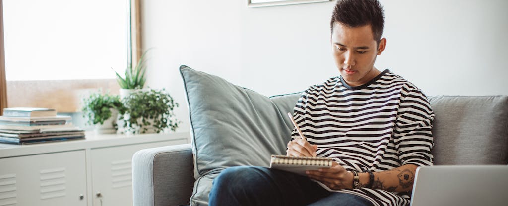 Young man sitting on sofa in his apartment, writing on a notepad