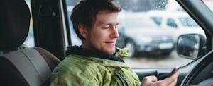 Young man wearing a green rain jacket sitting inside his car and checking his phone to see if his insurance covers windshield cracks