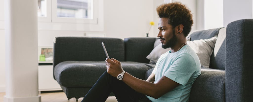 Young man sitting on floor at home with digital tablet, looking up how to set up direct deposit