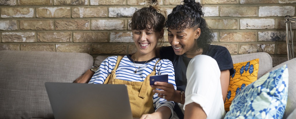 two young women on their couch at home shopping with their debit card on a laptop