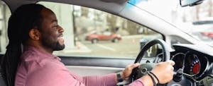 A man in a pink long sleeve shirt smiles while driving his new car.