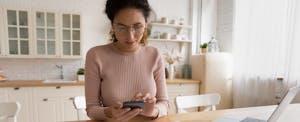 A woman seated at her kitchen table uses a calculator and laptop to create a budget.