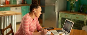 A woman seated at a table with a notepad and a calculator uses her new MacBook to take an online course.