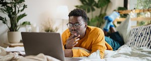 A man lying on a bed uses a laptop to look into a share-secured loan.