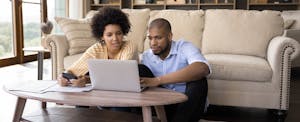 A couple seated on the floor in front of a coffee table use a laptop to submit a joint loan application.