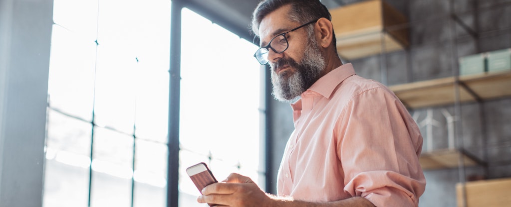 A bearded man sits near a large window and uses his smartphone to research a PNC checking account.