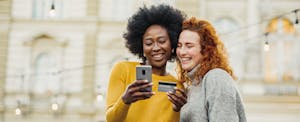 united-quest-card Two smiling women stand side by side looking at a smartphone, while one holds her United Quest Card in her other hand.