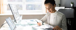 A woman seated at a desk with both a laptop and desktop computer uses a calculator while assessing an investment opportunity.