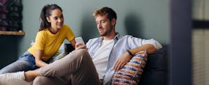 A man seated on a sofa looks thoughtfully at his smartphone while showing the screen to his partner who is seated next to him.