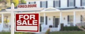 Windows and front porch of a large white home in the background, with a red and white for sale (forclosure) sign in the foreground