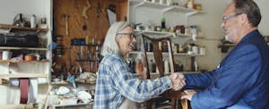 A furniture restorer stands next to a chair in their workshop and shakes hands with a client after a price negotiation.