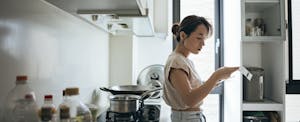 Young woman in her kitchen, leaning against her stove as she reviews a grocery receipt