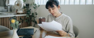 A focused woman seated at a table reviews financial documents while calculating annual income at home using a laptop.
