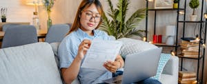 A woman sitting on a couch reviews a paper document in front of a laptop that is resting on her knees.
