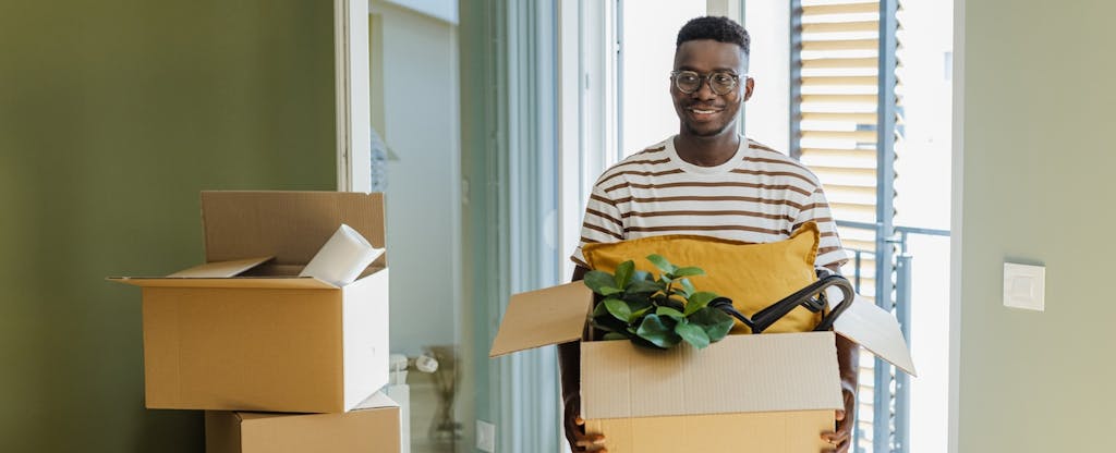 A man carries a cardboard box full of pillows and houseplants into a new apartment.