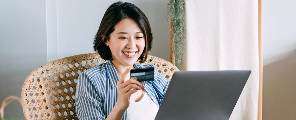 Smiling young Asian woman sitting on a rattan armchair in the living room at home, shopping online with laptop and making mobile payment with credit card. With some shopping bags next to her